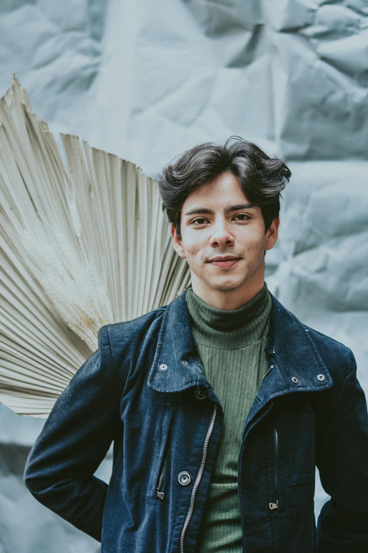Studio portrait of a young man in a jacket, standing in front of a dried palm leaf backdrop